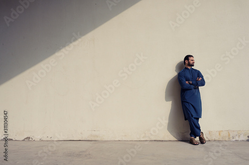 Young man with beard wearing blue long traditional cloth standing at wall wtih sunlight and shadow background, Isolated.