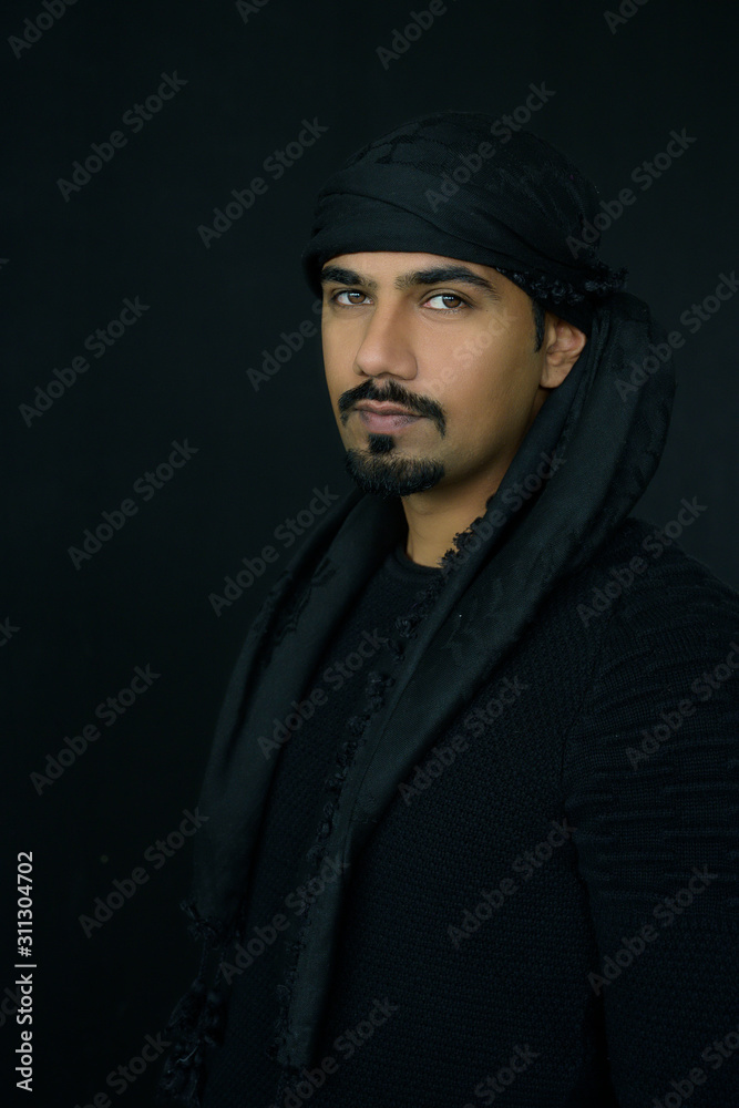 Handsome middle east man with beard wearing long black dress with turban standing in dark room background, Bahrain. Handsome middle east man with beard wearing long black dress with turban standing in dark room background, Bahrain.