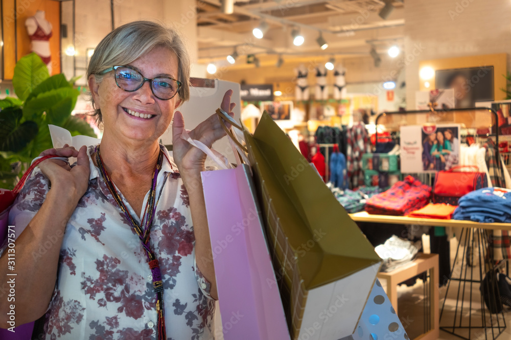 Front view of a senior woman smiling and satisfied with purchases in a mall. Gray hair and eyeglasses. Loaded with shopping bags Front view of a senior woman smiling and satisfied with purchases in a mall. Gray hair and eyeglasses. Loaded with shopping bags