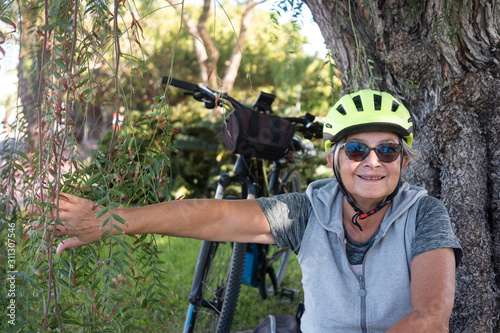 An attractive senior woman cyclist with yellow helmet have a break in the park under a big plant.. Moment of relaxation for a caucasian people