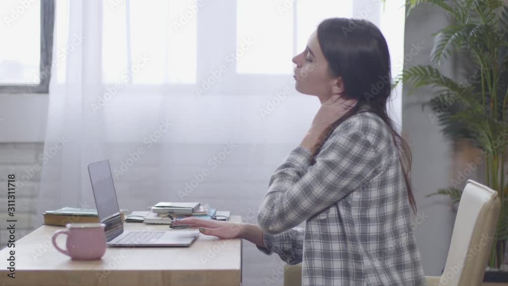 Young brunette Caucasian woman having ache in back while typing on laptop keyboard. Confident female freelancer exercising as sitting at the table. Distant work, incorrect sitting position. Young brunette Caucasian woman having ache in back while typing on laptop keyboard. Confident female freelancer exercising as sitting at the table. Distant work, incorrect sitting position.
