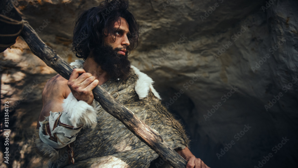 Primeval Caveman Wearing Animal Skin Holds Stone Hammer Comes out of Cave and Looks Around Prehistoric Landscape, Ready to Hunt Animal Prey. Neanderthal Going to Hunt in a Jungle. Low Angle Shot