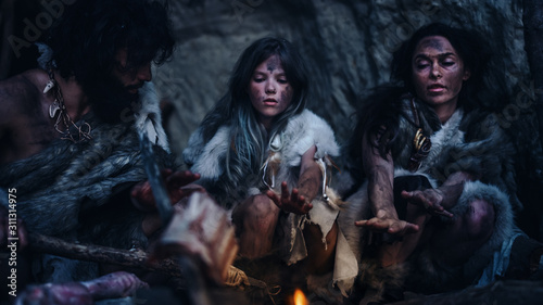 Tribe of Prehistoric Hunter-Gatherers Wearing Animal Skins Live in Cave at Night. Neanderthal Family Trying to Get Warm at the Bonfire, Holding Hands over Fire, Cooking Food. Shot with a Warm Filter.