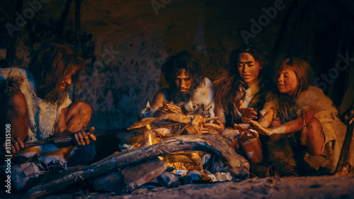 Neanderthal or Homo Sapiens Family Cooking Animal Meat over Bonfire and then Eating it. Tribe of Prehistoric Hunter-Gatherers Wearing Animal Skins Grilling and Eating Meat in Cave at Night