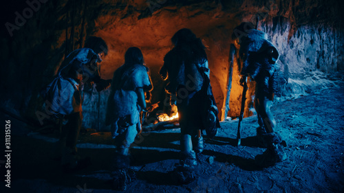Tribe of Prehistoric Hunter-Gatherers Wearing Animal Skins Live in a Cave at Night. Neanderthal or Homo Sapiens Family Trying to Get Warm at the Bonfire, Holding Hands over Fire. Back View