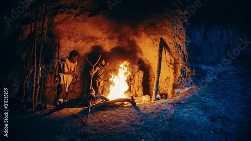 Tableau sur toile Tribe of Prehistoric Hunter-Gatherers Wearing Animal Skins Stand Around Bonfire Outside of Cave at Night