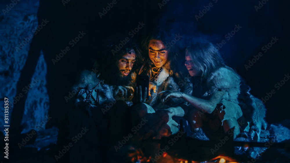 Tribe of Prehistoric, Primitive Hunter-Gatherers Wearing Animal Skins Use Smartphone in a Cave at Night. Neanderthal / Homo Sapiens Family Browsing Internet on Mobile Phone