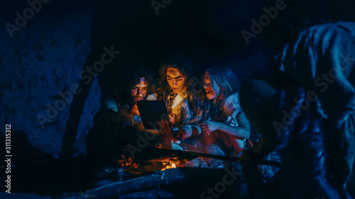 Tribe of Prehistoric  Primitive Hunter-Gatherers Wearing Animal Skins Use Digital Tablet Computer in a Cave at Night. Neanderthal or Homo Sapiens Family Browsing Internet  Watching Videos  Streaming 