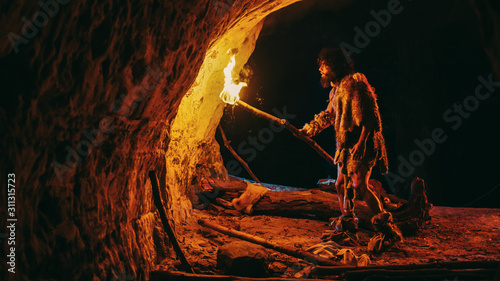 Canvastavla Primeval Caveman Wearing Animal Skin Exploring Cave At Night, Holding Torch with Fire Looking at Drawings on the Walls at Night