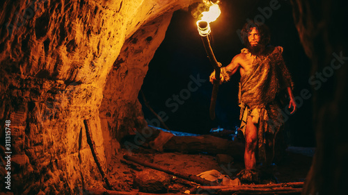 Primeval Caveman Wearing Animal Skin Exploring Cave At Night, Holding Torch with Fire Looking at Drawings on the Walls at Night. Neanderthal Searching Safe Place to Spend the Night photo