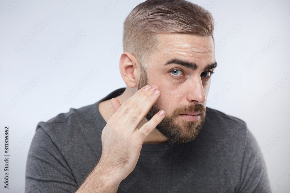 Horizontal close up portrait shot of handsome bearded man massaging moisturising face cream into his skin looking at camera, white background