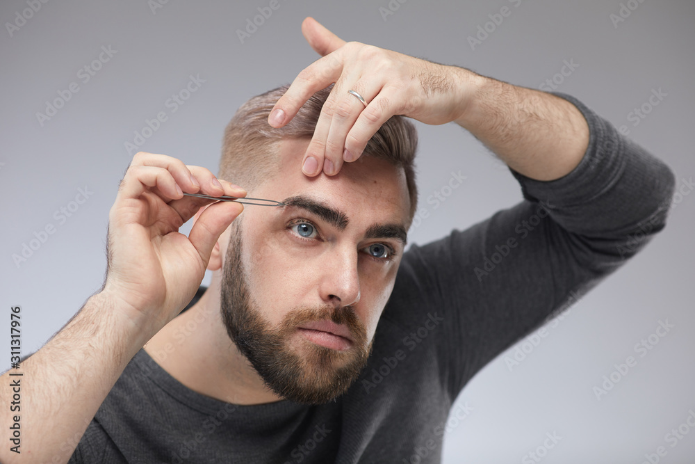 Obraz premium Studio portrait of stylish young man standing against gray wall background shaping his eyebrows using tweezers
