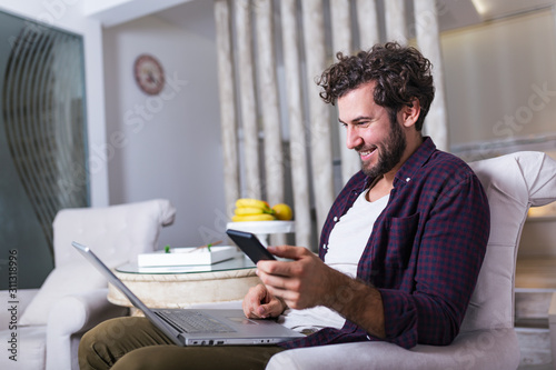 Successful entrepreneur smiling in satisfaction as he checks information on his laptop computer while working in a home office. Young man relaxing on the sofa with a laptop and mobile phone in hands