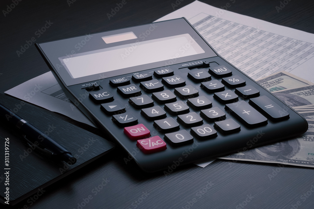 Black accounting calculator, notepad, pen, report with a numerical table and several hundred dollar bills on a dark wooden table. Stock trading, calculation of taxes, profits, and incomes concept Black accounting calculator, notepad, pen, report with a numerical table and several hundred dollar bills on a dark wooden table. Stock trading, calculation of taxes, profits, and incomes concept