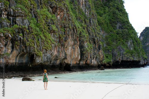 A girl with red hair in a green dress stands on the seashore and looks into the distance. A sea of ​​amazing azure color. The girl stands next to a huge rocky back to the viewer.