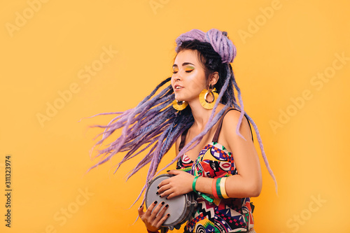 Hipster woman with violet dreadlocks and colored clothes playing on ethical mini drum on yellow background © Peakstock
