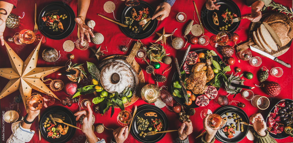 Friends celebrating Christmas. Flat-lay of people eating and talking over festive table with red cloth with champagne, roasted chicken, bundt cake, fruits, decorations, top view, wide composition Friends celebrating Christmas. Flat-lay of people eating and talking over festive table with red cloth with champagne, roasted chicken, bundt cake, fruits, decorations, top view, wide composition