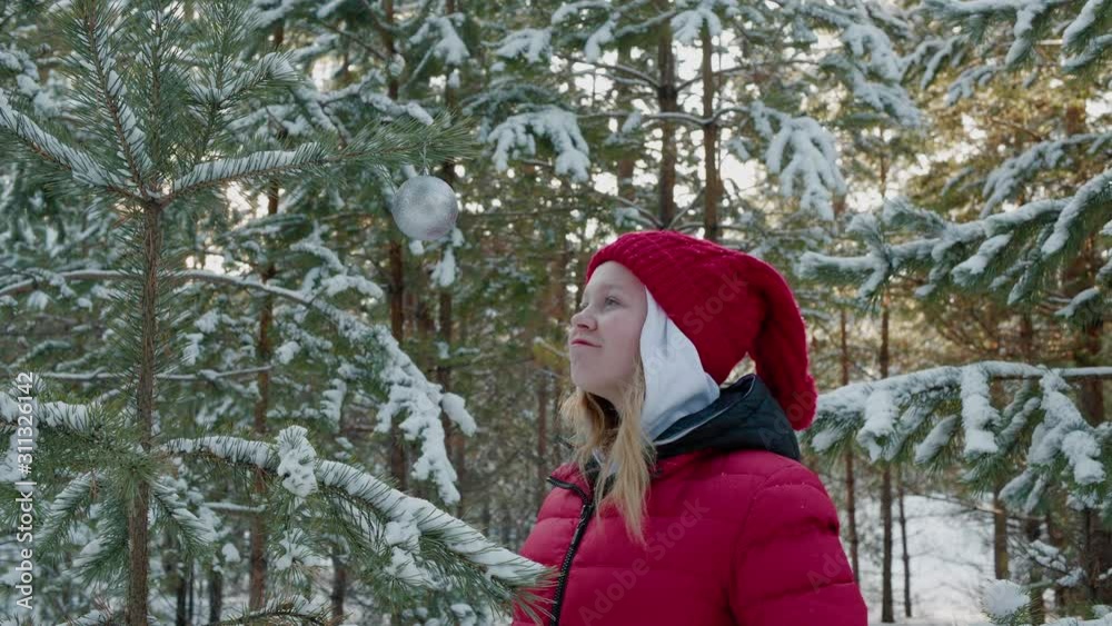 Young girl teenager playing with decoration ball on Christmas tree in snowy forest at winter walk. Happy girl touching silver ball hanging on New Year tree in winter forest. Young girl teenager playing with decoration ball on Christmas tree in snowy forest at winter walk. Happy girl touching silver ball hanging on New Year tree in winter forest.