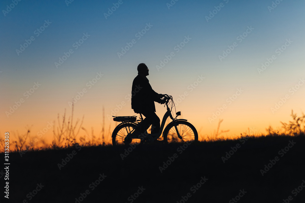 Obraz premium Male cyclist on the e-bike or electric bicycle on the sunset background riding up the hill. Silhouette of the old man in profile. Active pension. Travel. Sport.