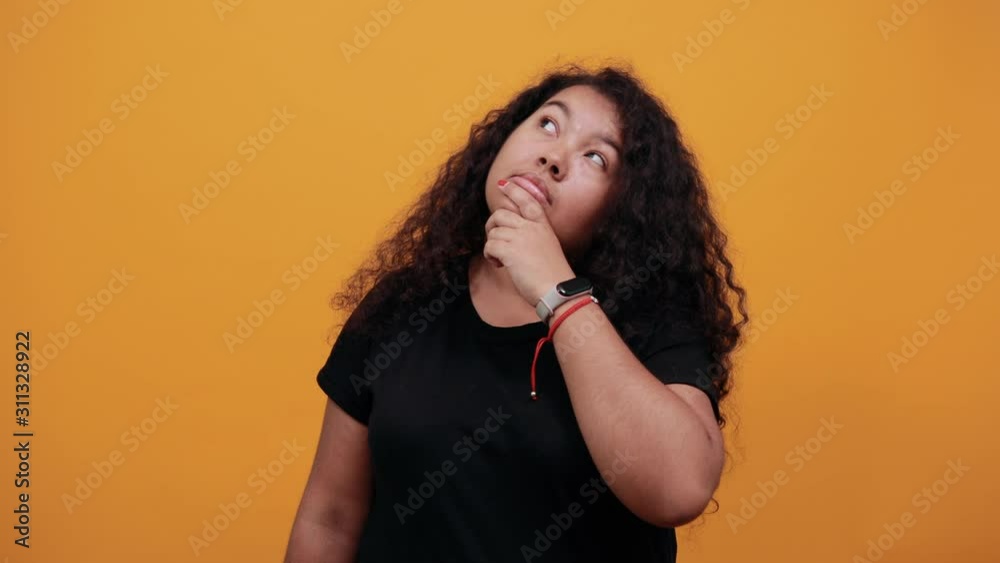 Strict afro-american young woman with overweight keeping finger on mouth, looking up, thinking, over isolated orange background wearing fashion black shirt. People lifestyle concept. Strict afro-american young woman with overweight keeping finger on mouth, looking up, thinking, over isolated orange background wearing fashion black shirt. People lifestyle concept.
