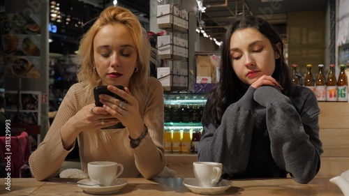 Two young beautiful women in cafe use watch smartphone chatting social media while anothe girlfriend boring drinking tea photo