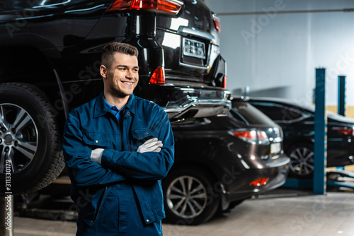 young, smiling mechanic standing with crossed arms and looking away near modern cars in workshop
