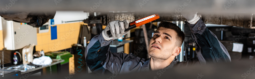 Fototapeta premium panoramic shot of mechanic inspecting car bottom with flashlight