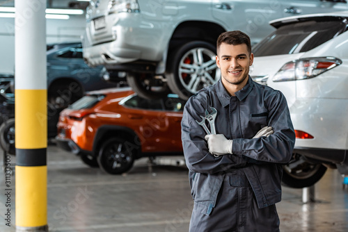 smiling mechanic looking at camera while standing with crossed arms and holding wrenches