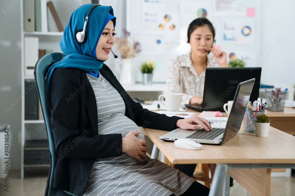 Fototapeta premium Cute pregnant muslim woman talking by headset in office. maternity businesswoman in suit having conversation online solving customer problem in workplace. asian chinese female colleague in back