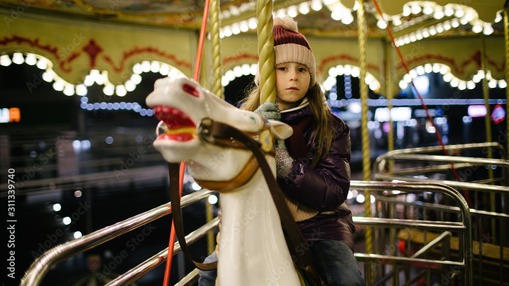 Sad kid girl riding a carousel on a white horse at night