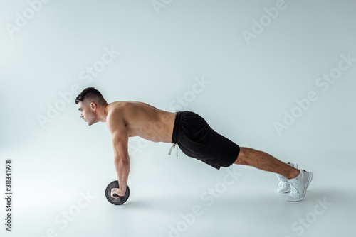 side view of sexy muscular bodybuilder with bare torso exercising with ab wheel on grey background