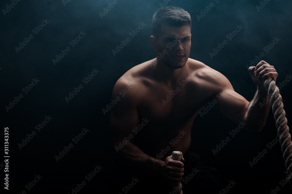 sexy muscular bodybuilder with bare torso excising with battle rope on black background with smoke