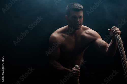 sexy muscular bodybuilder with bare torso excising with battle rope on black background with smoke