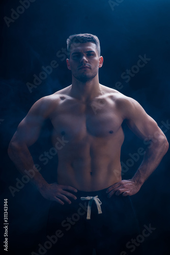 sexy muscular bodybuilder with bare torso posing with hands on hips on black background with smoke