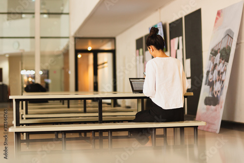 Back view of young businesswoman using cell phone with open laptop on table.