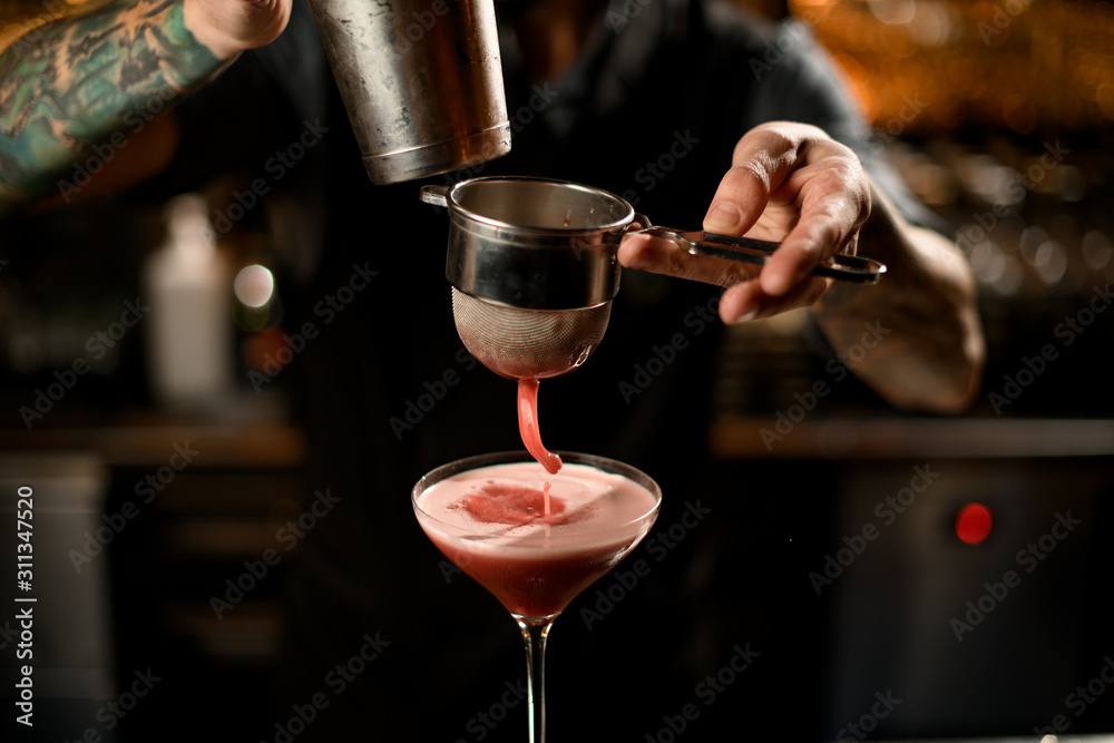 Bartender pouring a alcoholic drink from the steel shaker to the glass through the sieve