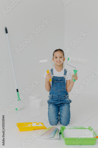 Happy little girl in denim overalls stands on knees, holds paint roller, surrounded with trays with paint, poses in white room, has happy expression, helps parents with house improvement and painting