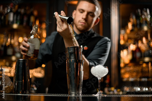 Attractive male bartender pouring a liqour from the jigger to a professional steel shaker