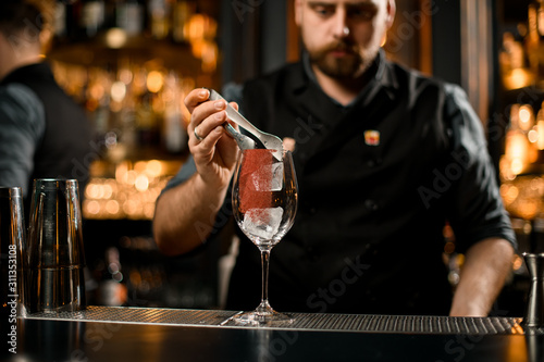 Male bartender putting an ice cubes with tweezers to the empty cocktail glass with a red spice decor