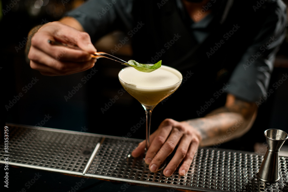 Bartender decorated the yellow creamy color alcoholic cocktail drink with a green leaf with tweezers