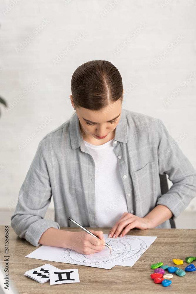 Astrologer drawing natal chart beside cards and stones with zodiac signs on table Astrologer drawing natal chart beside cards and stones with zodiac signs on table