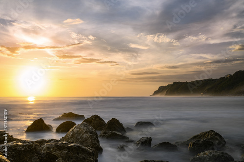 (Selective focus) Stunning view of a rocky coast bathed by a smooth silky sea during sunset. Melasti Beach with its cliff in the distance, South Bali, Indonesia.