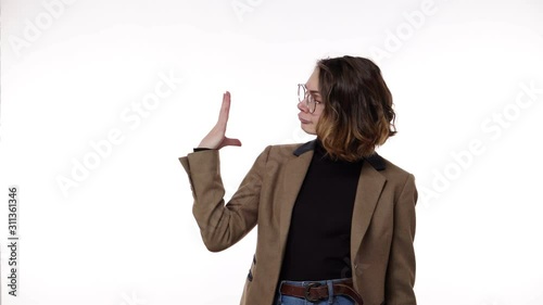 Annoyed pretty woman standing in front the camera isolated on a white background and showing blahblah gesture, gossips. Irritated, emotional. Wearing brown, retro jacket, jeans and stylish glasses photo