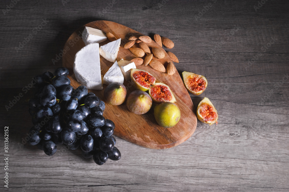 Still life cheese plate on a wooden cutting board lies sliced cheese with mold almonds in the shell and sliced figs next to a bunch of black grapes. Delicious and healthy food concept