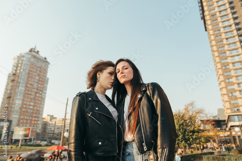 Fashion portrait of two girls in street clothes standing on grass on cityscape background and posing at camera. Portrait of attractive models in leather jackets on the street.