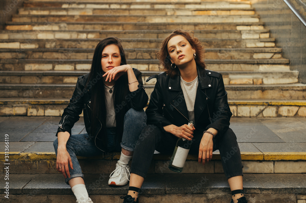 Portrait of two girls models in stylish casual clothes sitting on stairs with bottle of wine in hand posing at camera with serious face. Two girlfriends are sitting on the stairs in the underground Portrait of two girls models in stylish casual clothes sitting on stairs with bottle of wine in hand posing at camera with serious face. Two girlfriends are sitting on the stairs in the underground