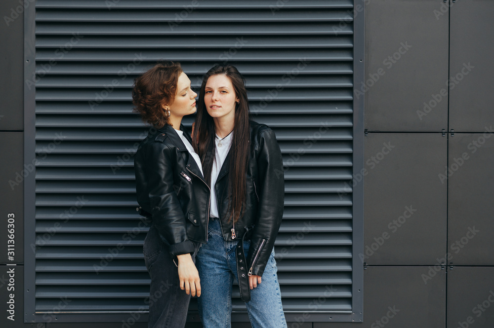 Portrait of two sexy girls in leather jackets, standing on a dark wall background and posing at camera. Pair of lesbians stands on black background. Pride concept. Beautiful girlfriend on background.