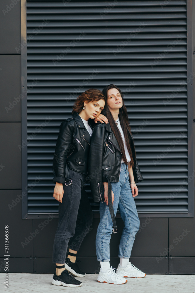 Portrait of two beautiful models on a dark wall background, wearing stylish casual clothes, posing for the camera.Two buttresses are standing outside on a dark background and looking at the camera Portrait of two beautiful models on a dark wall background, wearing stylish casual clothes, posing for the camera.Two buttresses are standing outside on a dark background and looking at the camera