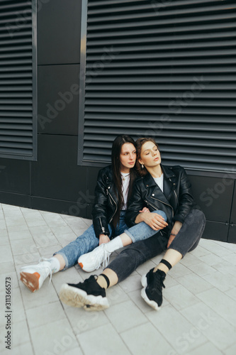 Fashionable photo of 2 stylish girls in casual clothes,wear leather jacket,sitting on the ground against a dark wall and looking away.Street portrait of two girlfriends sitting on a pavement.Vertical © bodnarphoto