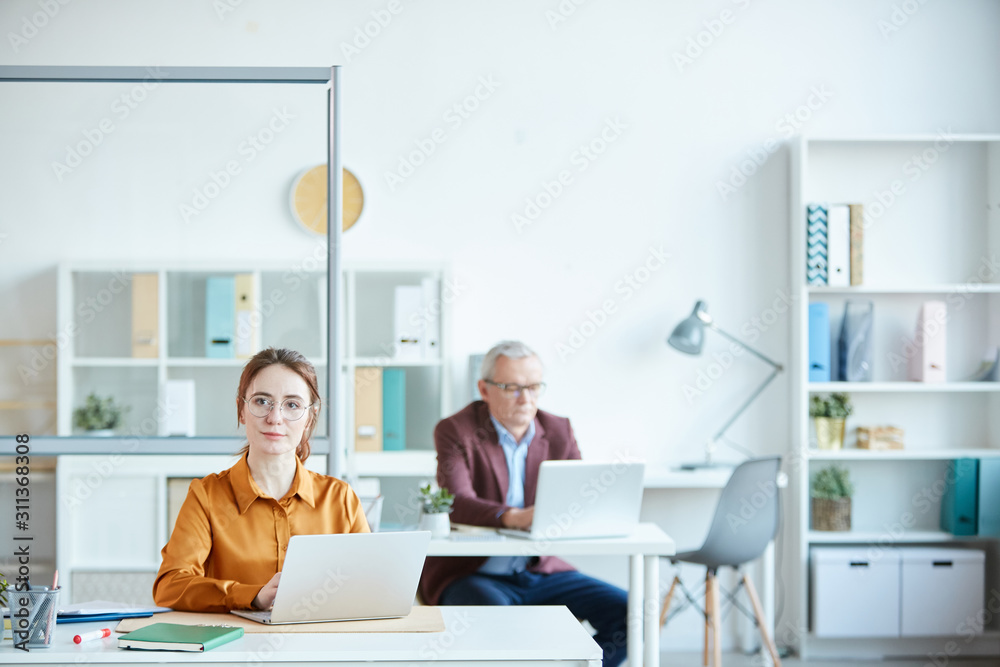 Portrait of young businesswoman in eyeglasses working on laptop and looking at camera with mature man working in the background Portrait of young businesswoman in eyeglasses working on laptop and looking at camera with mature man working in the background